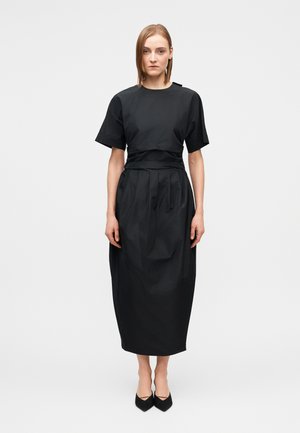 Naine musta lühikeste varrukatega midi-kleit plekkidega ja vööga vöökohal, seisab valge ühetoonilise tausta ees, näoga ettepoole.