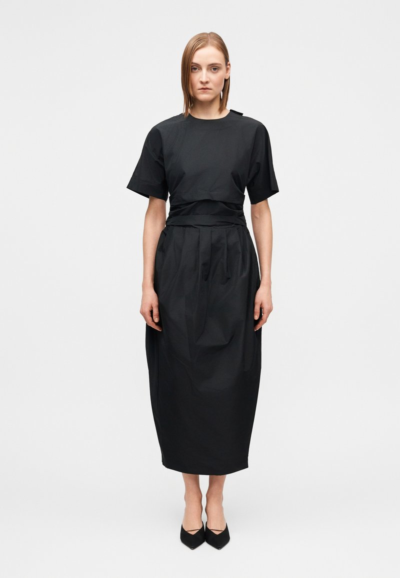 Woman wearing a black short-sleeve midi dress with pleats and a waist tie, standing against a plain white background, facing forward.