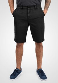 Homme portant un short noir arrivant au genou, des baskets bleu marine, et une chemise gris avec les mains dans les poches, se tenant devant un fond blanc uni.