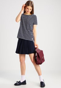 Camicia a maniche corte a righe blu navy e bianche abbinata a una gonna plissettata blu navy, calze bianche e mocassini neri, mentre tiene una borsa a tracolla bordeaux.