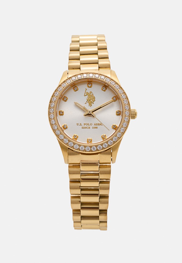 Montre dorée avec un cadran rond, un cadran argenté scintillant, des accents en cristal et un bracelet en maillons métalliques dorés. Présente un logo à 12 heures.