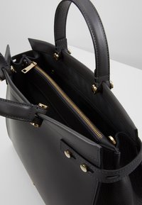 Musta nahkalaukku, jossa on kaksoiskahvat, kultainen vetoketjuniho sisällä ja kultanväriset varaosat, asetettuna vaalealle pinnalle.