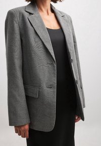 Nainen, joka on pukeutunut harmaaseen herringbone-bleiseriin mustan hihattoman mekon päällä, seisoo vaaleaa taustaa vasten.