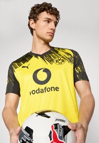 Gul fodboldtrøje med sorte detaljer, der har et diagonalt grafisk design, logo og sponsortekst. Spilleren holder en sort-hvid fodbold.