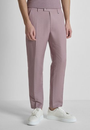 Parte inferiore del corpo con pantaloni slim fit color malva, una maglietta abbinata e scarpe da ginnastica bianche con lacci su uno sfondo uniforme.