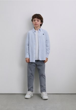 Camisa de cuadros azul claro con botones sobre una camiseta blanca, combinada con pantalones grises y zapatillas blancas. De pie contra un fondo neutro.