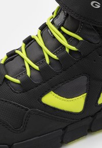 Chaussure de sport noire avec des lacets et des accents vert fluo. Tige en synthétique texturé avec un design découpé, semelle en caoutchouc épaisse pour l'adhérence.