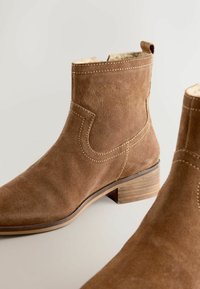 Ghete maro din piele întoarsă, până la gleznă, cu vârf rotunjit, cusături decorative, toc scăzut și o buclă pentru tragere la spate. Căptușeală interioară moale vizibilă.