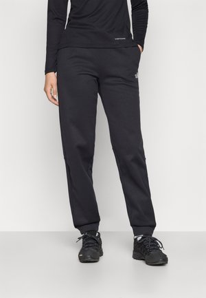 Osoba ubrana w czarną bluzkę z długim rękawem, czarne spodnie dresowe i czarne trampki stojąca na jasnym, jednolitym tle.