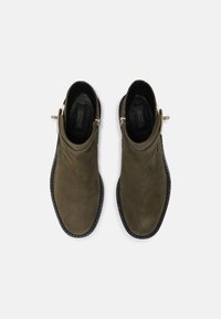 Une paire de bottines en daim olive foncé avec des semelles noires et des bouclettes dorées sur les côtés, vue de dessus sur un fond blanc.