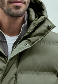 Oliven grøn polstret jakke med lynlås krave, der har sølv hardware og en tekstureret indvendig foring. Glat materiale med synlige sømme.
