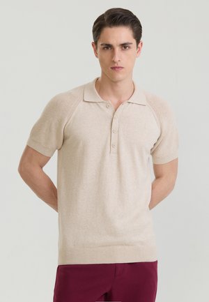 Joven vistiendo una camisa tipo polo de punto beige de manga corta y pantalones burdeos, de pie con las manos detrás de la espalda frente a un fondo liso.