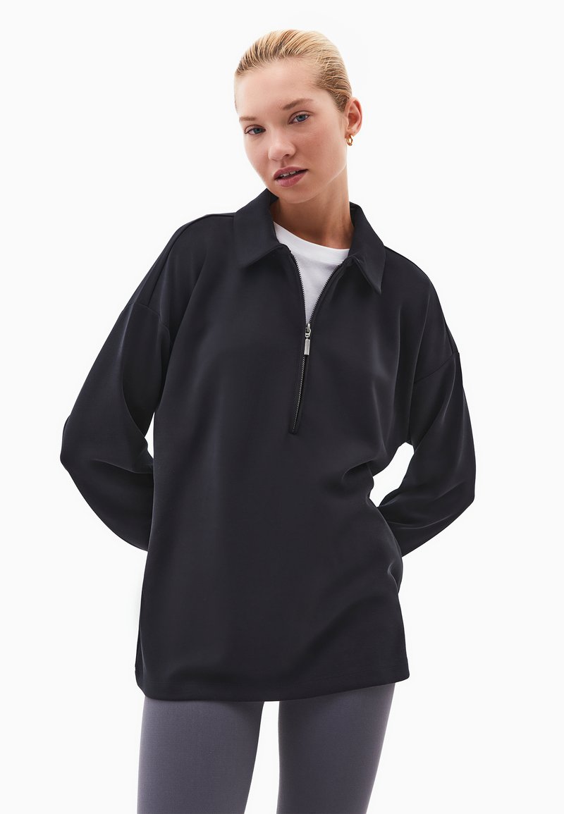 Černý pulovr se zipem a volným střihem, vyrobený z měkké látky, s límcem a dlouhými rukávy, vrstvený přes bílou košili.