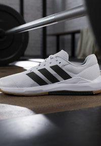 Chaussure de sport blanche avec des rayures noires posée sur le sol d'une salle de sport près d'une barre avec des poids en arrière-plan.