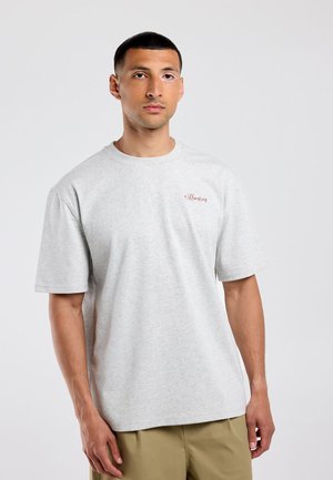 Man met kort haar die een lichtgrijs T-shirt draagt met kleine rode tekst op de linkerborst en beige broek, staand tegen een effen achtergrond.