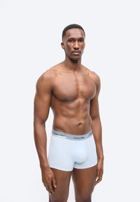 Urheilullinen mies seisoo yksivärisen taustan edessä valkoiset Calvin Klein -bokserit päällään, hieman vasemmalle päin kasvoillaan neutraali ilme.