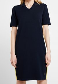 Granatowa sukienka z krótkimi rękawami, z dekoltem w kształcie litery V i żółtymi paskami po bokach. Wykonana z gładkiego, elastycznego materiału. Posiada logo w pobliżu dołu.