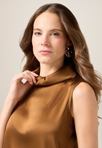 Bruine satijnen top met een gedrapeerde kraag, zachte textuur en mouwloos ontwerp. Model houdt de stof met één hand vast en toont de neklijn.