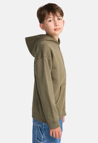 Ragazzo con capelli corti castani che indossa una felpa verde oliva con zip e jeans blu, in piedi di lato contro uno sfondo bianco semplice.