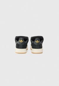 Melni zamšīgi sporta apavi ar gumijas zolēm, kuriem ir mīksta apkakle un zelta logotipa akcenti uz papēža. Dizainā iekļauts balts šuvums.