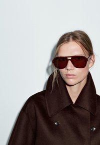 Brązowy, oversize'owy płaszcz z szerokim kołnierzem, z dwoma czarnymi guzikami. Modelka nosi duże, ciemnoczerwone, kątowe okulary przeciwsłoneczne z przyciemnionymi soczewkami.