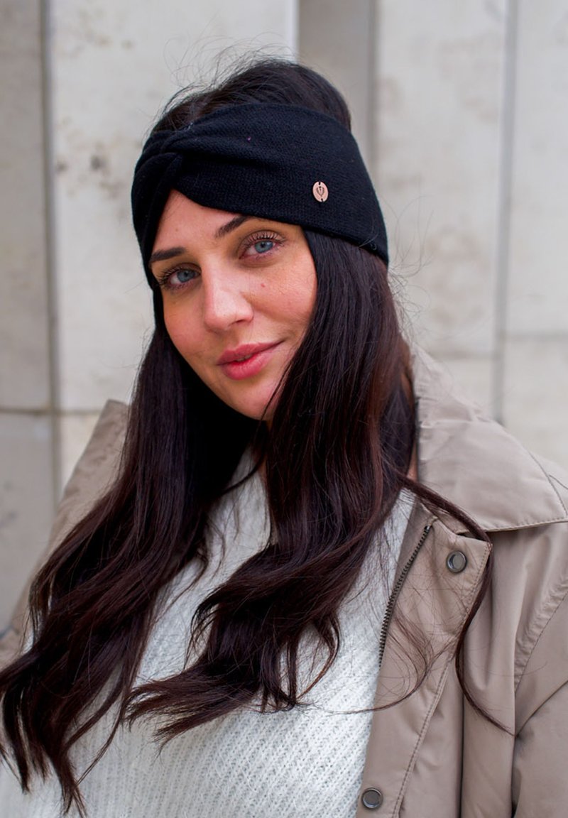Bandeau en tricot noir avec un design torsadé et un petit logo en métal, porté avec un léger pull et un manteau beige sur un fond uni.