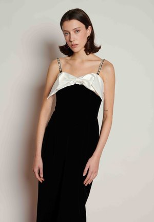 Νεαρή γυναίκα με κοντά καστανά μαλλιά, ντυμένη με μαύρο φόρεμα με λευκή λεπτομέρεια φιόγκου και διακοσμητικές λουρίδες με πολύτιμους λίθους, στέκεται μπροστά σε ένα ανοιχτό ουδέτερο φόντο.