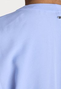 Gros plan sur le haut du dos d'une personne portant une chemise bleu clair avec un petit logo de marque métallique près de l'encolure.