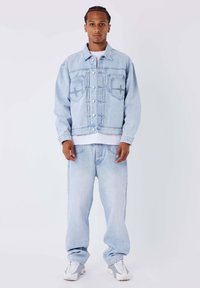 Giacca di jeans azzurro chiaro con chiusura a bottoni e due tasche sul petto, abbinata a jeans a gamba larga coordinati e sneakers bianche. Texture liscia.