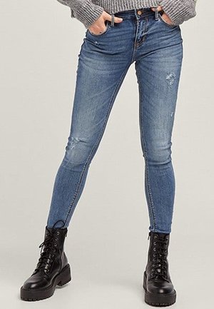 Slim-fit blå slidte jeans båret med sorte snøreløse kampstøvler og en grå sweater, hænder i lommerne.