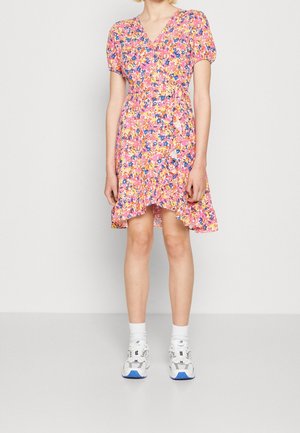 Kvinde iført en pink, orange og blå blomsteromslagkjole med flæser og pufærmer, kombineret med hvide ankelsokker og hvide sneakers.