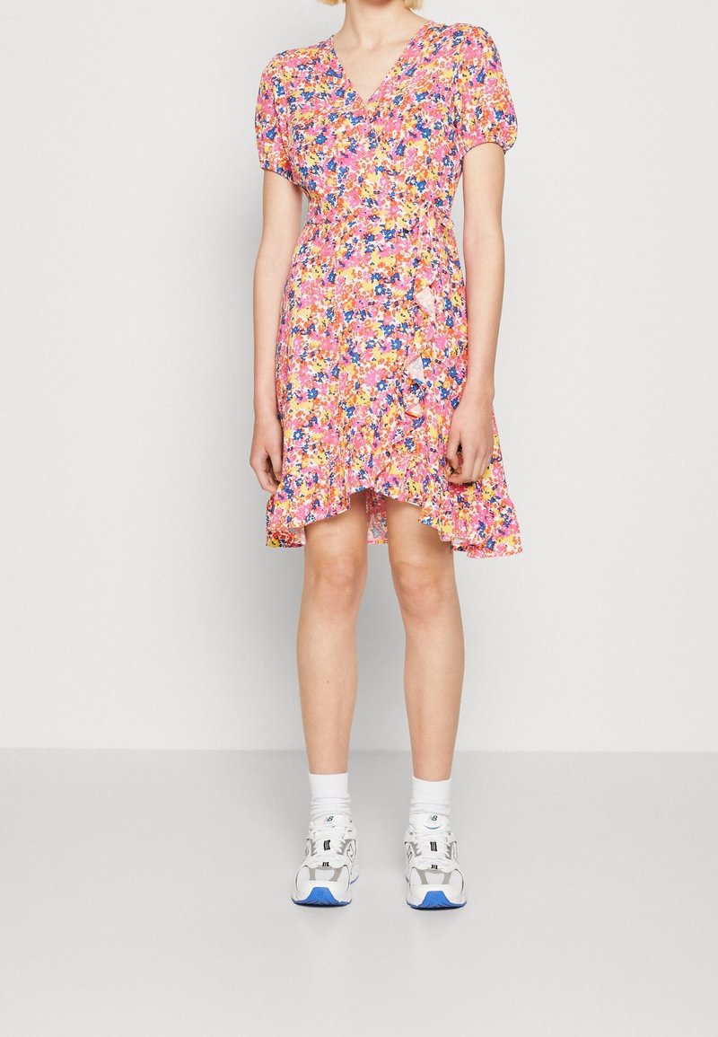 Femme portant une robe portefeuille fleurie rose, orange et bleue avec des volants et des manches bouffantes, associée à des chaussettes blanches chevilles et des baskets blanches.