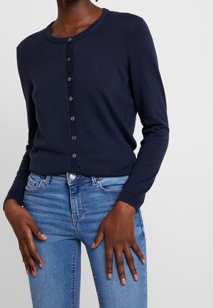 Vrouw draagt een donkerblauwe, gebuttonde cardigan en lichtblauwe spijkerbroek, handen rustend op de dijen, staand tegen een effen lichte achtergrond.