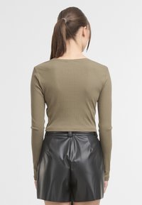 Žena s hnědými vlasy v culíku, oblečená v těsném olivově zeleném pleteném topu s dlouhým rukávem a černých kožených šortkách s vysokým pasem, zády k nám.