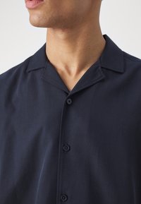 Camisa de botón azul marino con cuello muesca, mangas cortas y una textura suave. Presenta botones negros a lo largo de la tapeta delantera.