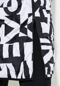 Vadderad väst med en djärv svart- och vit geometrisk mönstring, dragkedjestängning och en stilren yta. Svart tygbas med hög kontrastdesign.