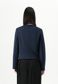 Personne avec des cheveux noirs raides portant un blazer bleu marine sur une jupe plissée noire, se tenant devant un fond blanc uni.