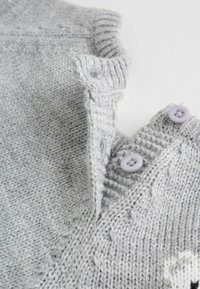 Close-up of light gray knitted fabric with ribbed collar, two sewn buttons, and a partial heart pattern in white yarn.