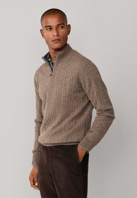 Bruine gebreide zip-up trui met ribbelstructuur en een hoge kraag, gelaagd over een blauw geruit overhemden, gedragen met donkere corduroy broek.