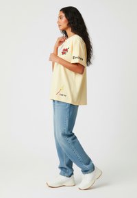 Lichtgele katoenen t-shirt met grafische ontwerpen, waaronder een hart en tekst. Gepaard met blauwe jeans en witte sneakers.