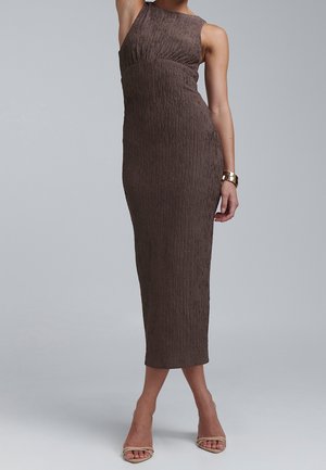 Femme portant une robe midi marron texturée sans manches, des talons ouverts et un bracelet en or, posant devant un fond gris clair uni.