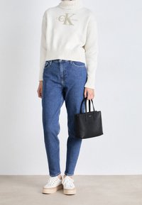 Weißer Strickpullover mit hohem Kragen und Logo, blaue Jeans, weiße Sneaker mit beige-farbener Plateausohle und eine kleine schwarze Handtasche.