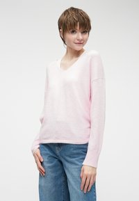 Svetlo roza pulover z v-izrezom in dolgimi rokavi, mehkega tekstura in rebrastimi manšeti. V kombinaciji z ohlapnimi modrimi kavbojkami.