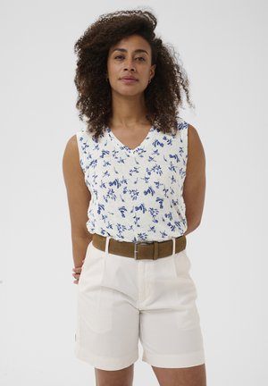 Femme aux cheveux bouclés portant un haut blanc sans manches avec un imprimé floral bleu, un short blanc et une ceinture marron, debout devant un fond uni.
