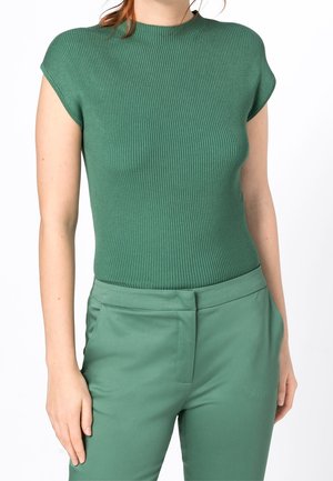 Vrouw draagt een geribd groen T-shirt met korte mouwen, ingestopt in een hoge tailles broek in het groen met een verborgen sluiting aan de voorkant.