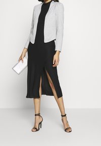 Černé šaty s bočním rozparkem, doplněné o texturovaný šedý blazer. Modelka drží stříbrnou kabelku a má na sobě černé sandály na podpatku.