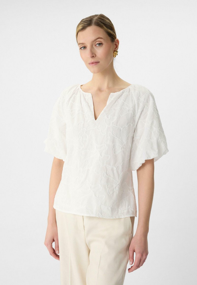 Weiße Bluse mit V-Ausschnitt und kurzen Puffärmeln, versehen mit floralem Stickerei und einem strukturierten Stoff. Getragen mit hellen Hosen.