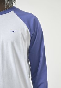 Weißes Raglan-T-Shirt mit lila Ärmeln, das ein kleines blaues Logo auf der Brust zeigt. Der Stoff sieht weich und glatt aus.