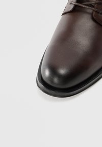 Barna bőr öltönycipő lekerekített orral, sima textúrával és fekete gumitalppal. Tonális fűzőkkel és minimalista dizájnnal rendelkezik.