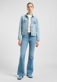 Giacca in denim azzurro chiaro con due tasche sul petto e bottoni, abbinata a jeans azzurri chiari a zampa d'elefante e scarpe da ginnastica coordinate.